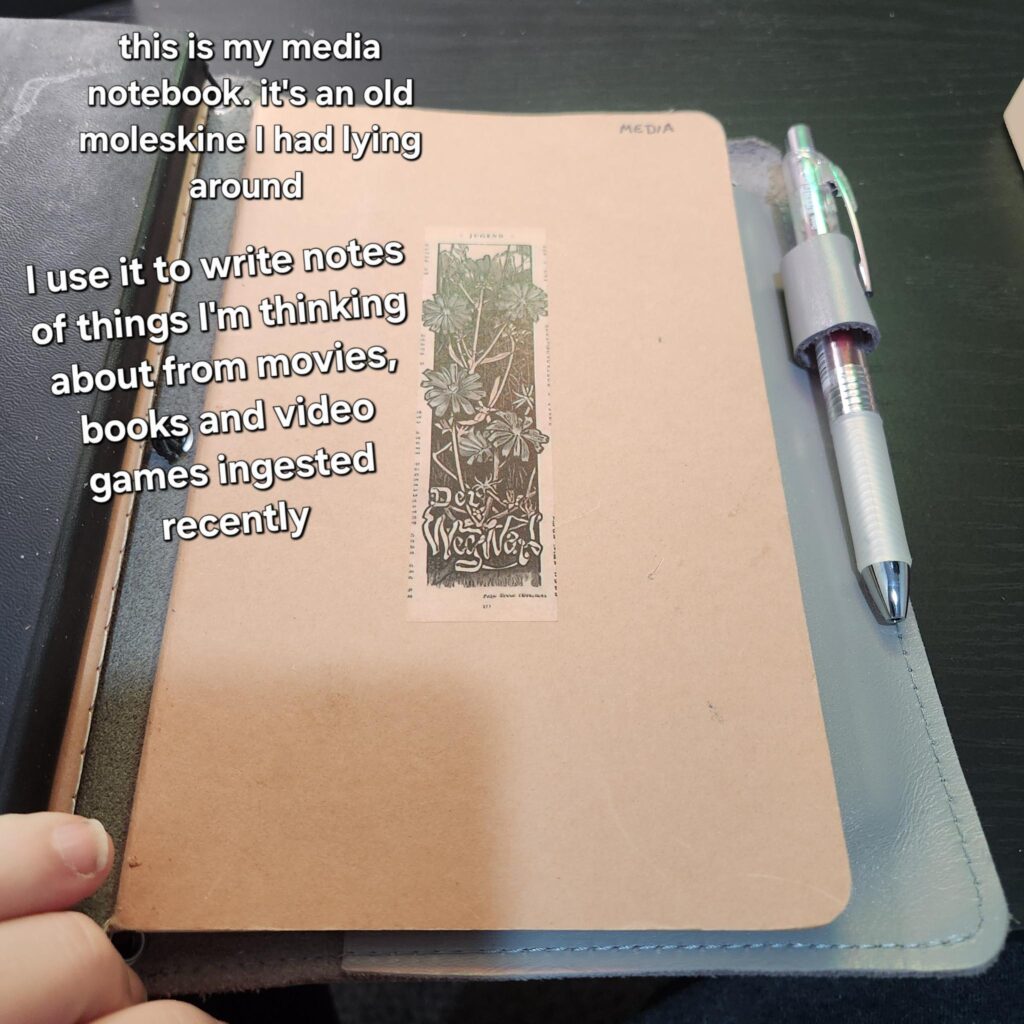Craft cardboard notebook with one sticker in the middle. Text: "This is my media notebook. it's an old moleskine I had laying around. I use it to write notes of things I'm thinking about from movies, books, and video games ingested recently.